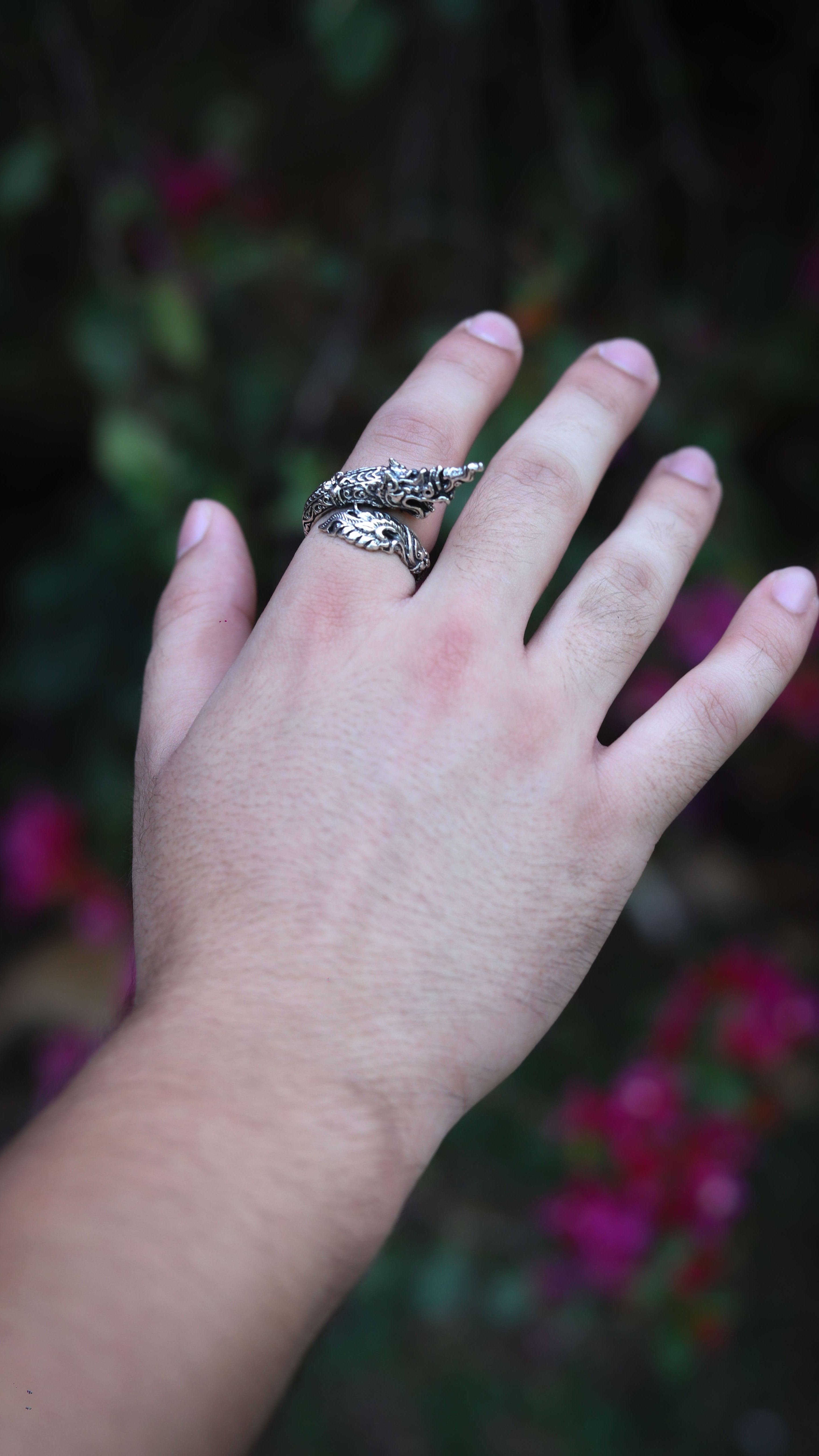 A silver-toned snake wrap ring worn on a person's ring finger, shown against a blurred outdoor background of green foliage and pink flowers.